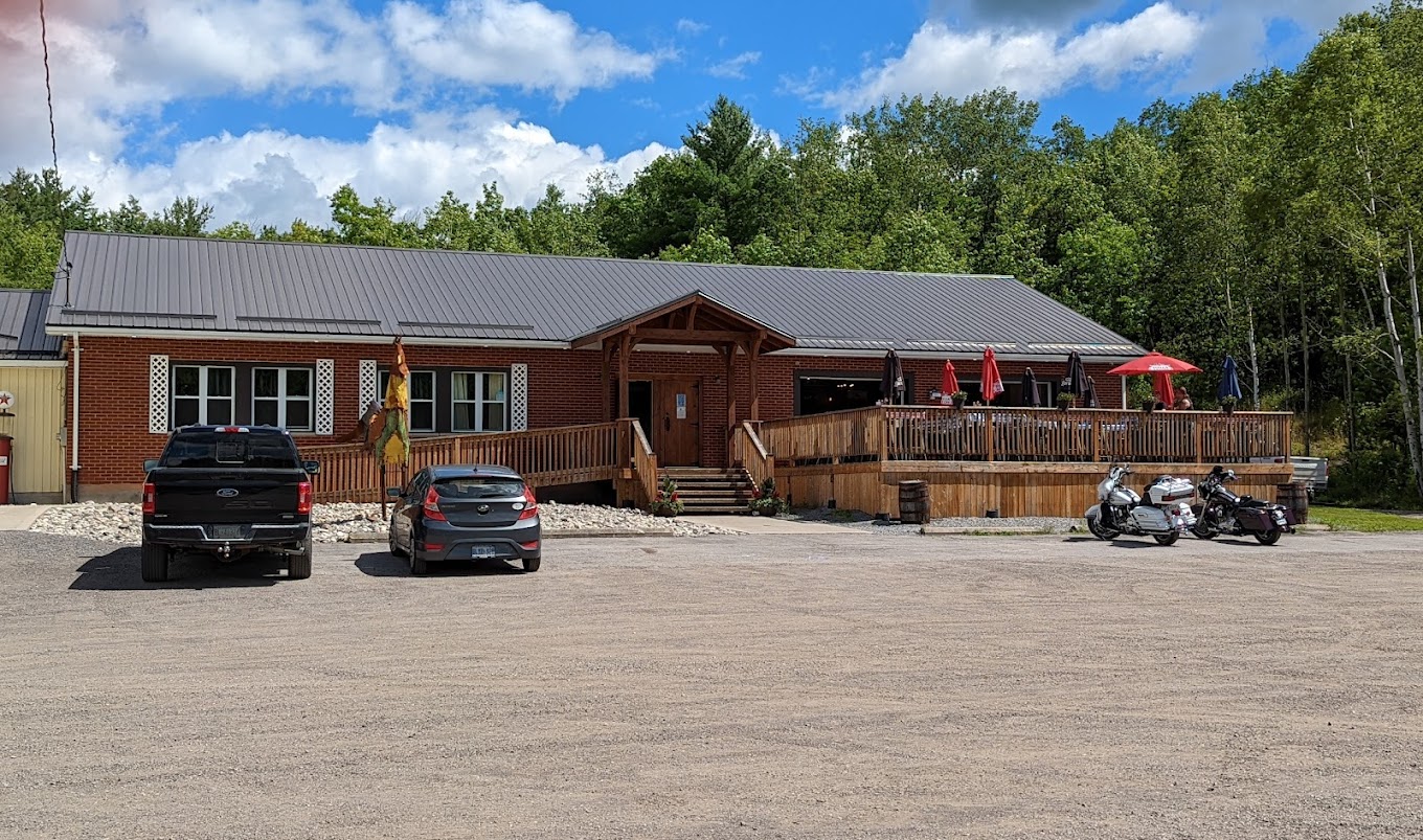 A single level building constructed of wood with two cars parked in front to the left and two motorcycles parked to the right. 