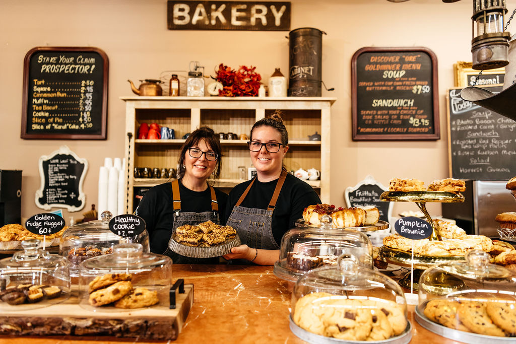 Two women smiling behind a counter with baked goods and pastries on it. They are holding a plate with food on it. Behind them is a wall with signs on it showing what's available on their menu.