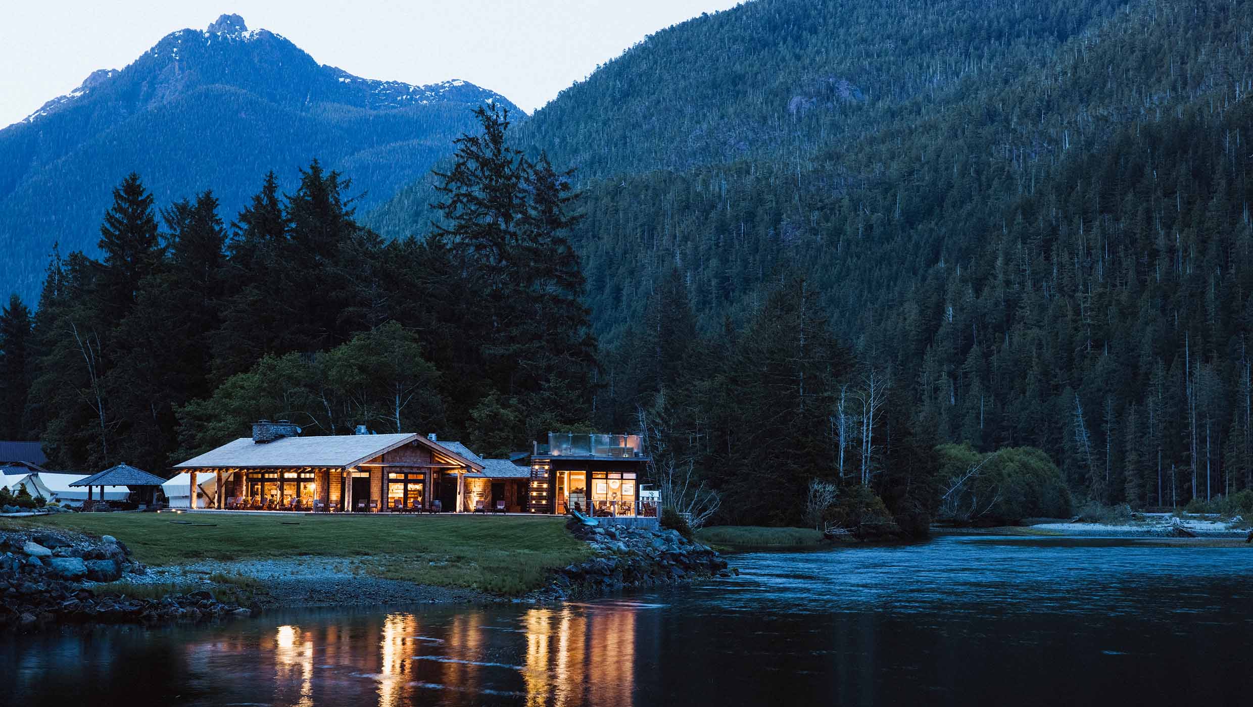 Clayoquot Wilderness lodge lit up in the evening, sitting on a river bank surrounded by spruce forest and mountains