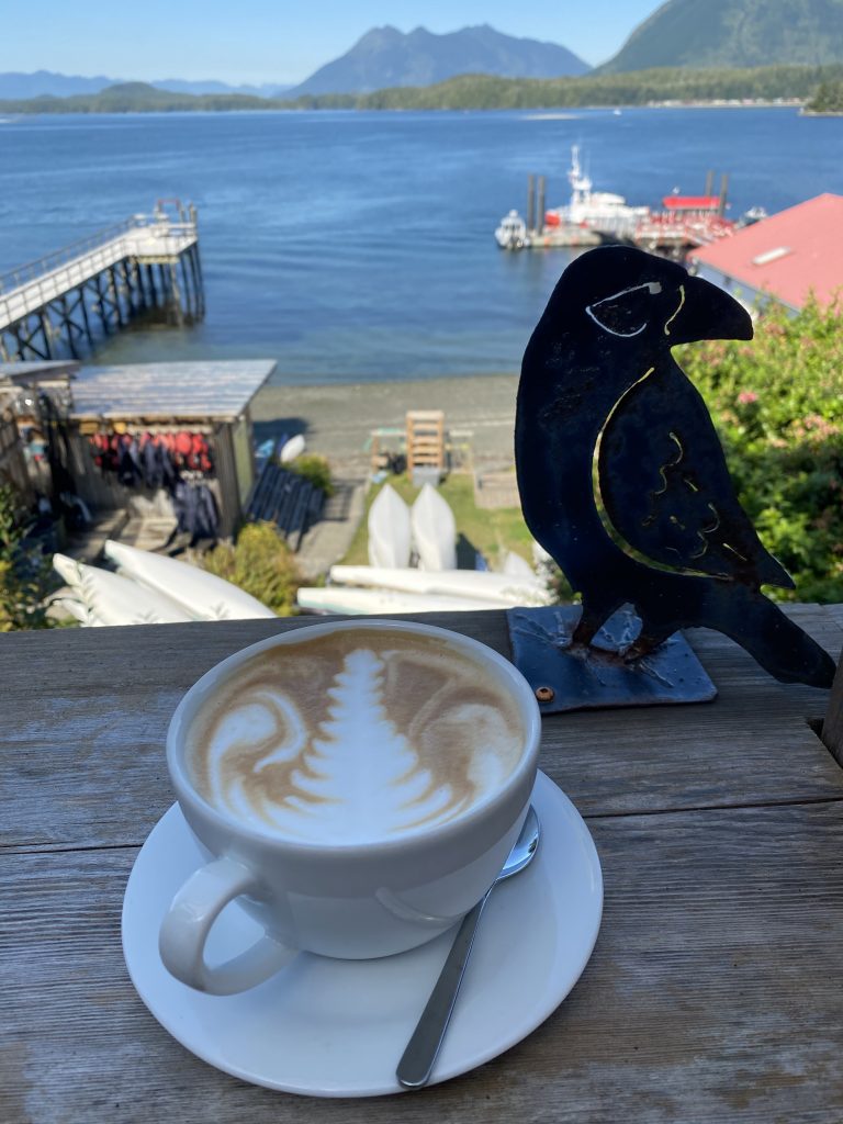 a latte with leaf expertly drawn in foam, in front of sea kayaks and the blue-grey waters, sky, and mountains of Tofino Harbour