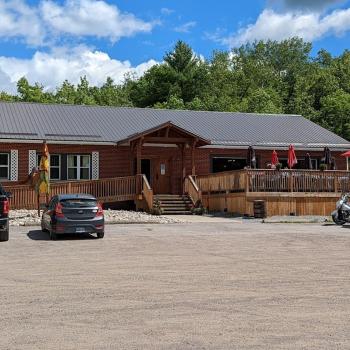 A single level building constructed of wood with two cars parked in front to the left and two motorcycles parked to the right. 