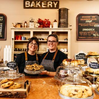 Two women smiling behind a counter with baked goods and pastries on it. They are holding a plate with food on it. Behind them is a wall with signs on it showing what's available on their menu.