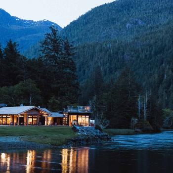 Clayoquot Wilderness lodge lit up in the evening, sitting on a river bank surrounded by spruce forest and mountains