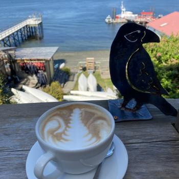 a latte with leaf expertly drawn in foam, in front of sea kayaks and the blue-grey waters, sky, and mountains of Tofino Harbour